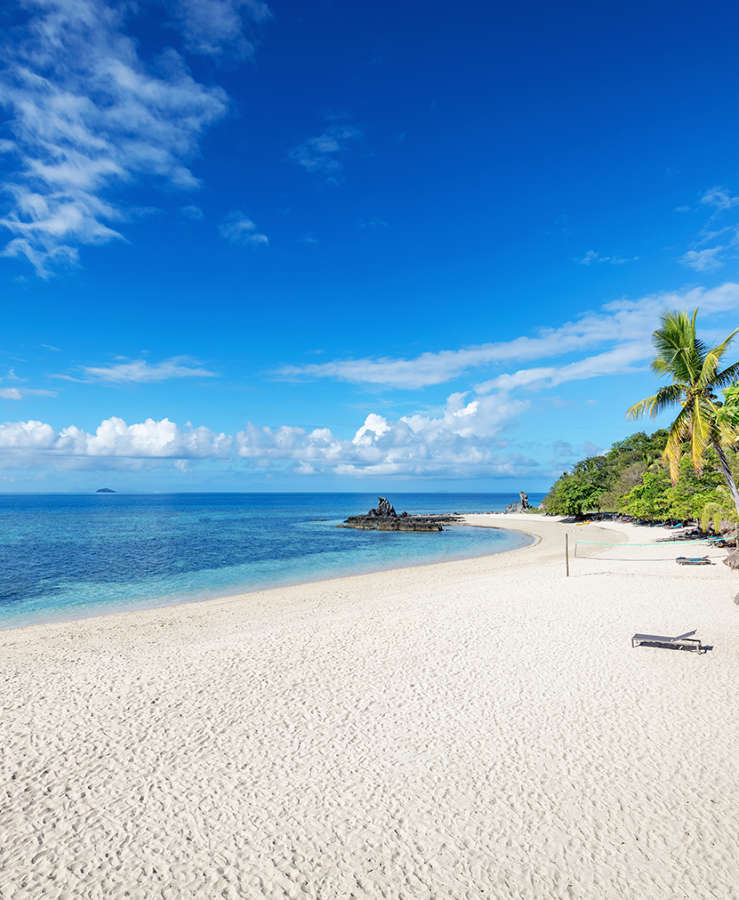 Beautiful beach with palm trees lounge chairs, beach volleyball net and beach huts on Castaway Island under blue summer sky. Mamanuca Islands, Fiji, Melanesia, South Pacific Ocean