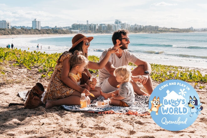 Family enjoying a beach picnic, Mooloolaba. Bluey's World Queensland logo.